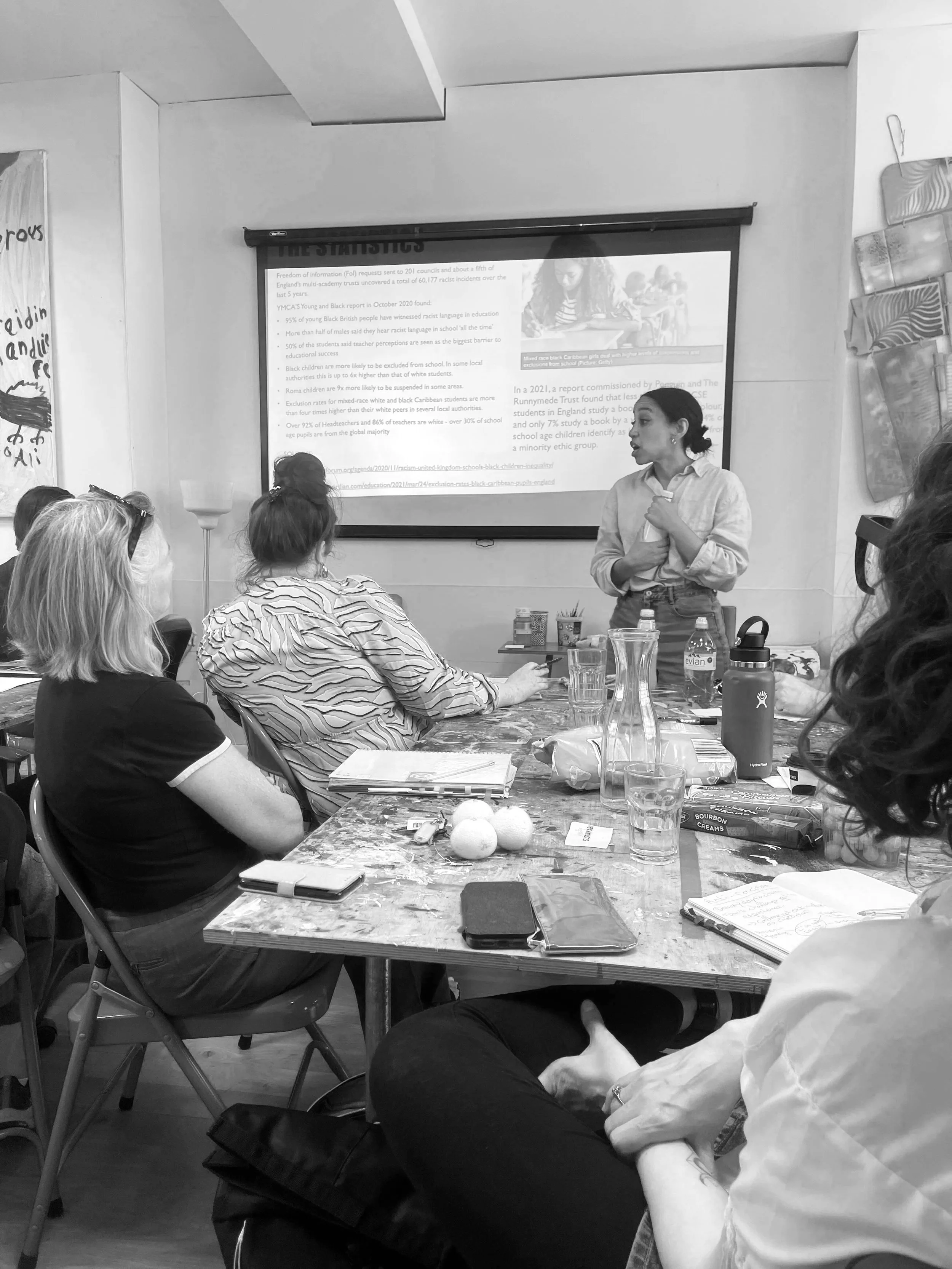A woman standing and speaking to a group of women seated at a table in a classroom or workshop setting, with a presentation slide projected on the wall behind her.