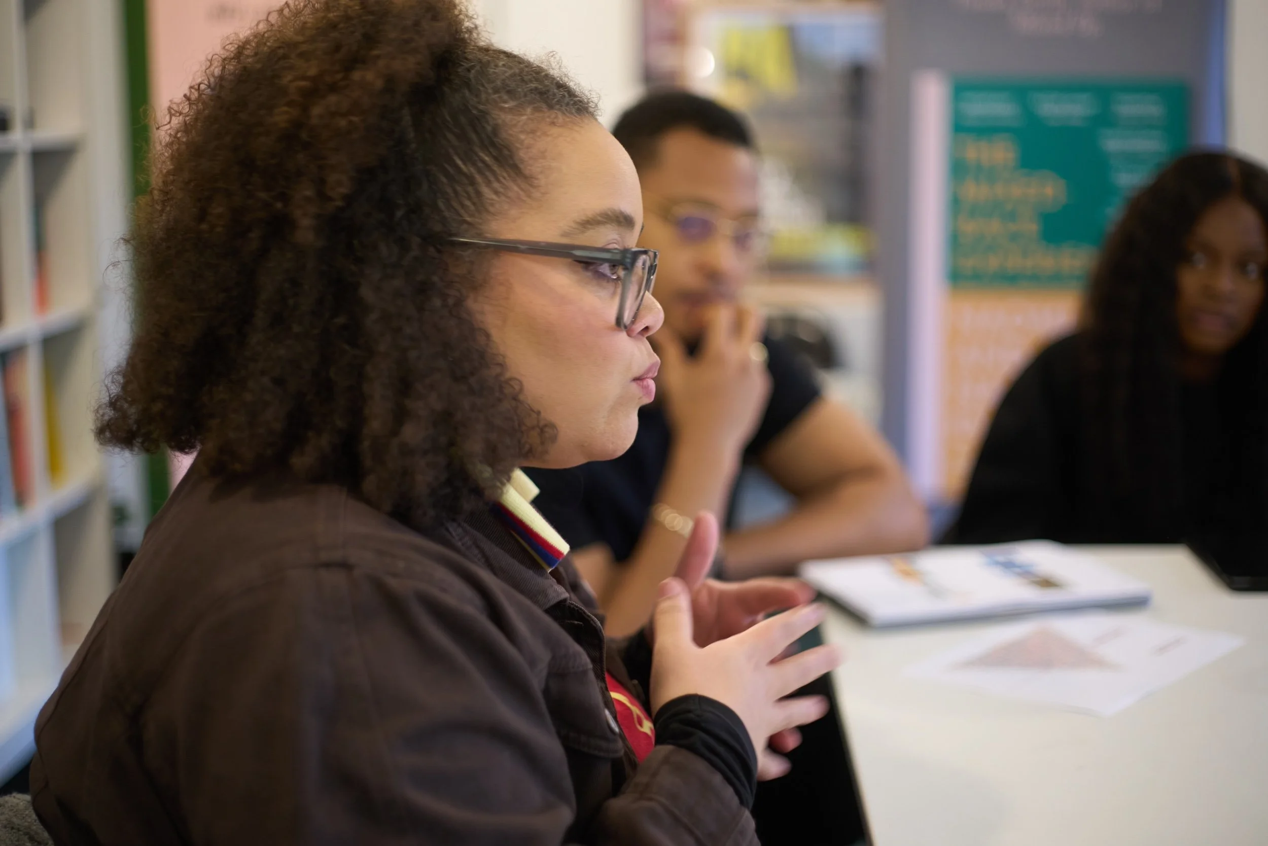 A woman with curly hair and glasses speaking while seated at a table in a group setting, with two other women listening in the background.