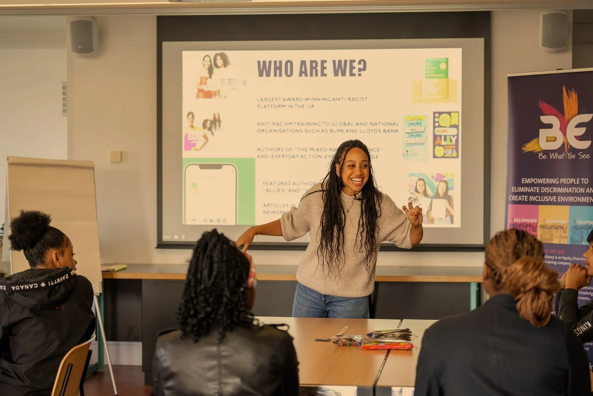 A woman smiling and speaking in front of a classroom with children. She is standing near a large screen displaying a presentation titled ‘Who Are We?’ about an anti-racism organization. Several children are seated, listening and looking at her.