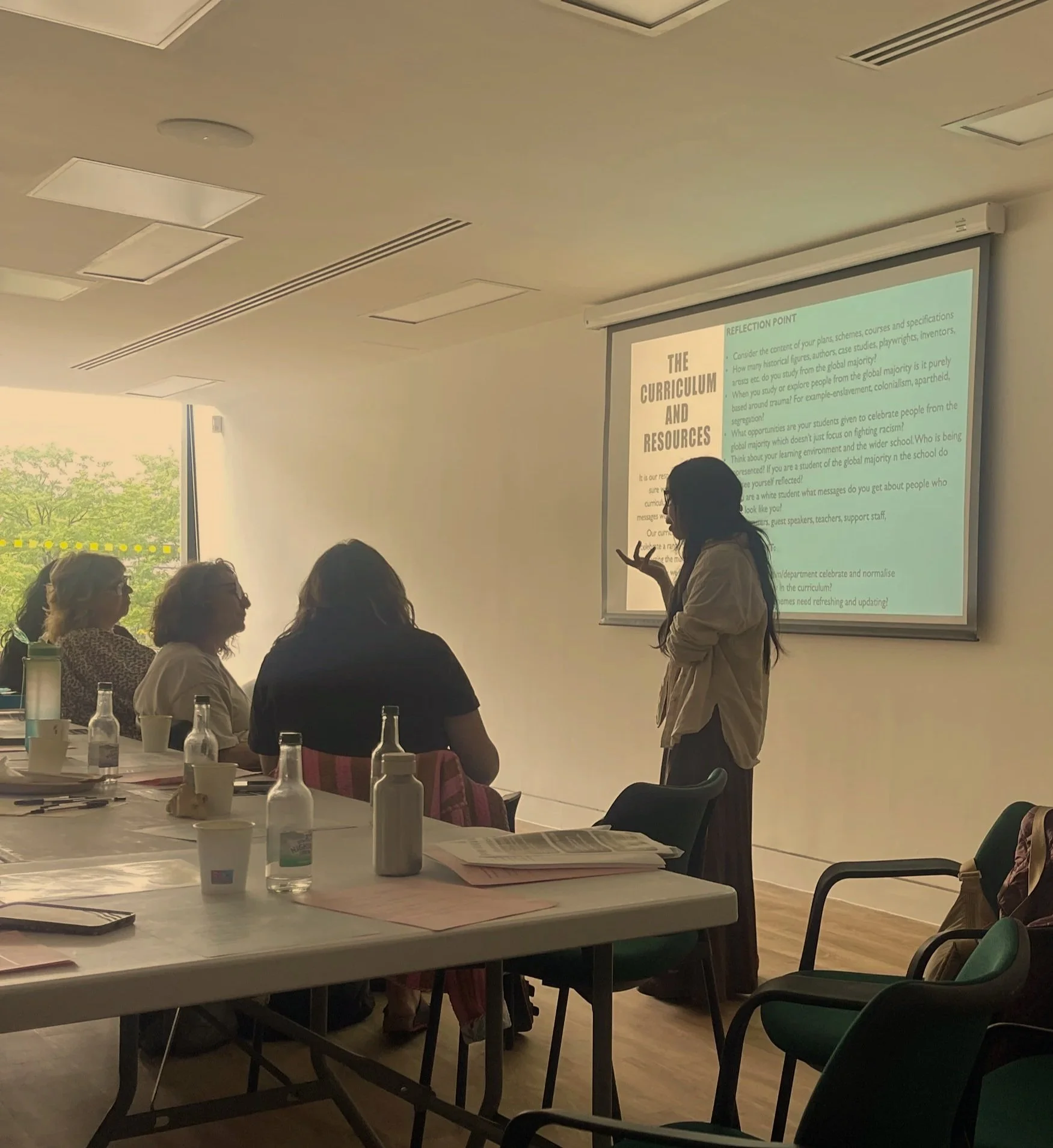 A woman giving a presentation to a seated audience in a conference room with a large window, white walls, and a projection screen displaying slides.