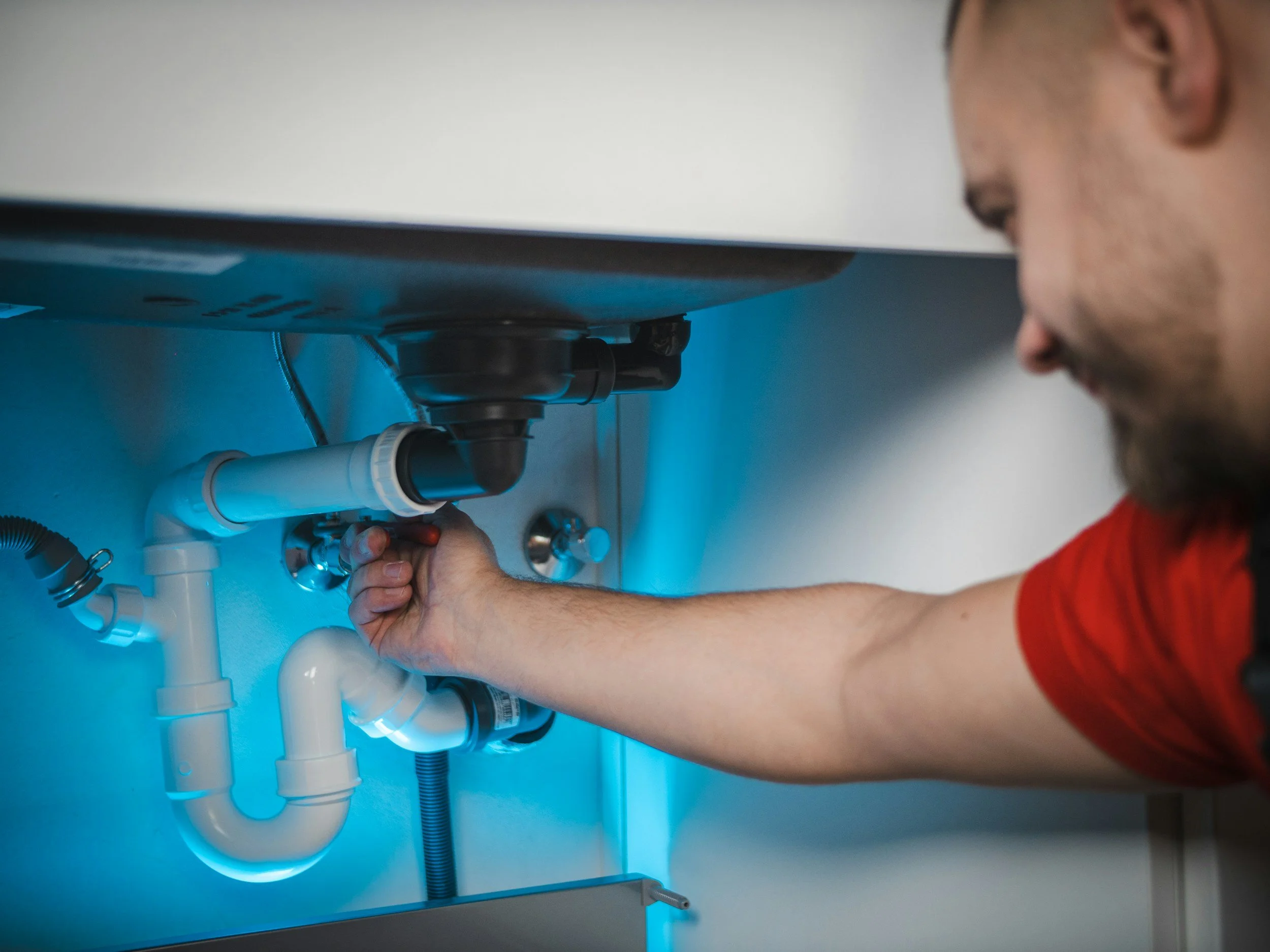 A man in a red shirt working under a sink, adjusting or inspecting the plumbing pipes.