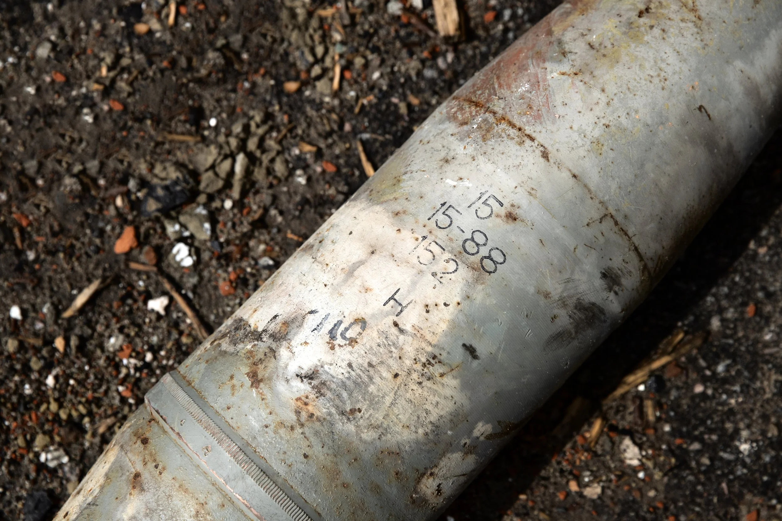 Close-up of an old, rusted cylindrical metal pipe lying on dirt ground, with markings and numerical stamps on its surface.