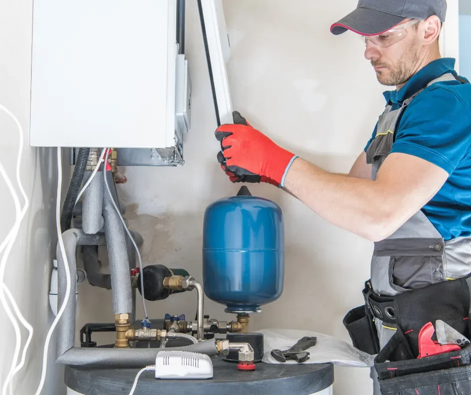 Technician installing or repairing a boiler or water heater, wearing safety glasses and red gloves, in a utility room.