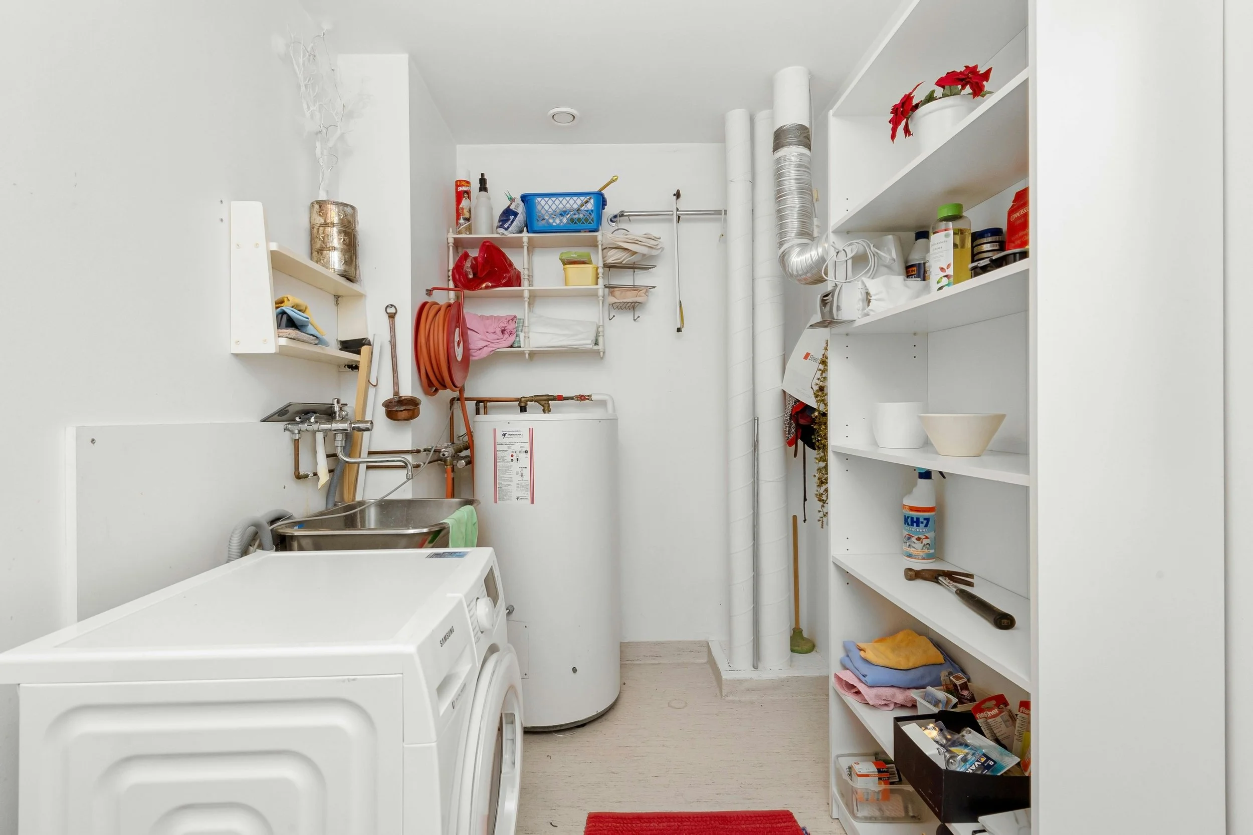 Laundry room with washing machine, utility sink, shelving units holding cleaning supplies, towels, and tools, and a red rug on a light-colored floor.