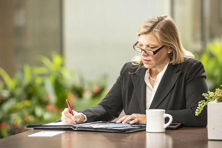 Businesswoman writing about marketing strategy in a notebook at a desk with a white coffee mug and plants in the background.