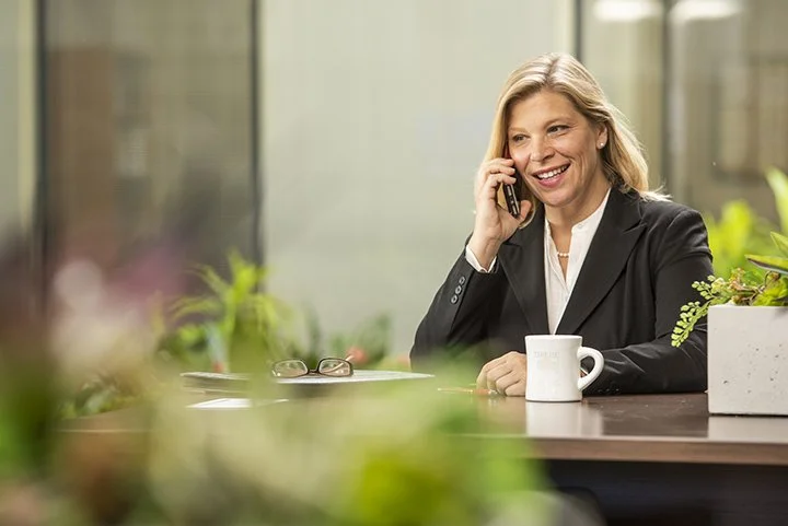 A woman in a black blazer talking on a phone about marketing strategy while sitting at a desk in a bright office setting.