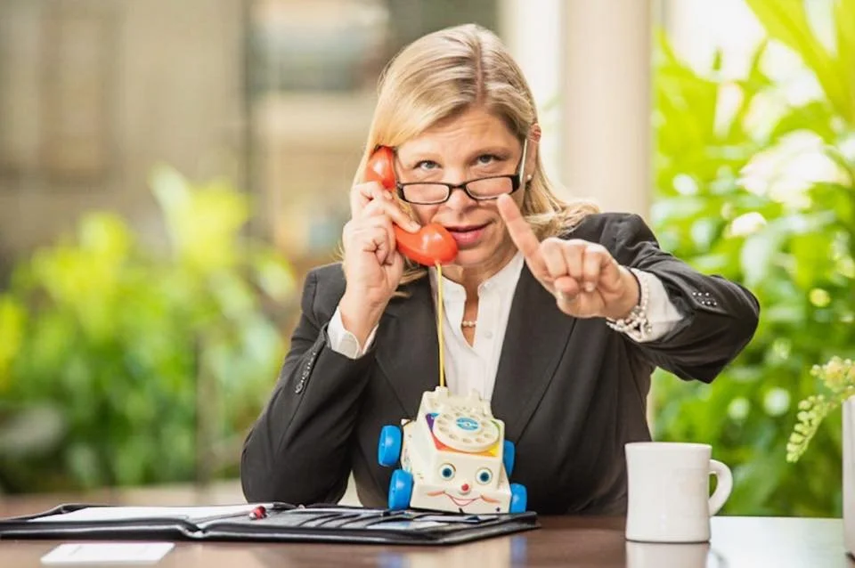 A woman with blonde hair, wearing glasses and a black blazer, talks on a vintage toy telephone, pointing her finger. She is sitting at a desk with a coffee mug and a notepad.