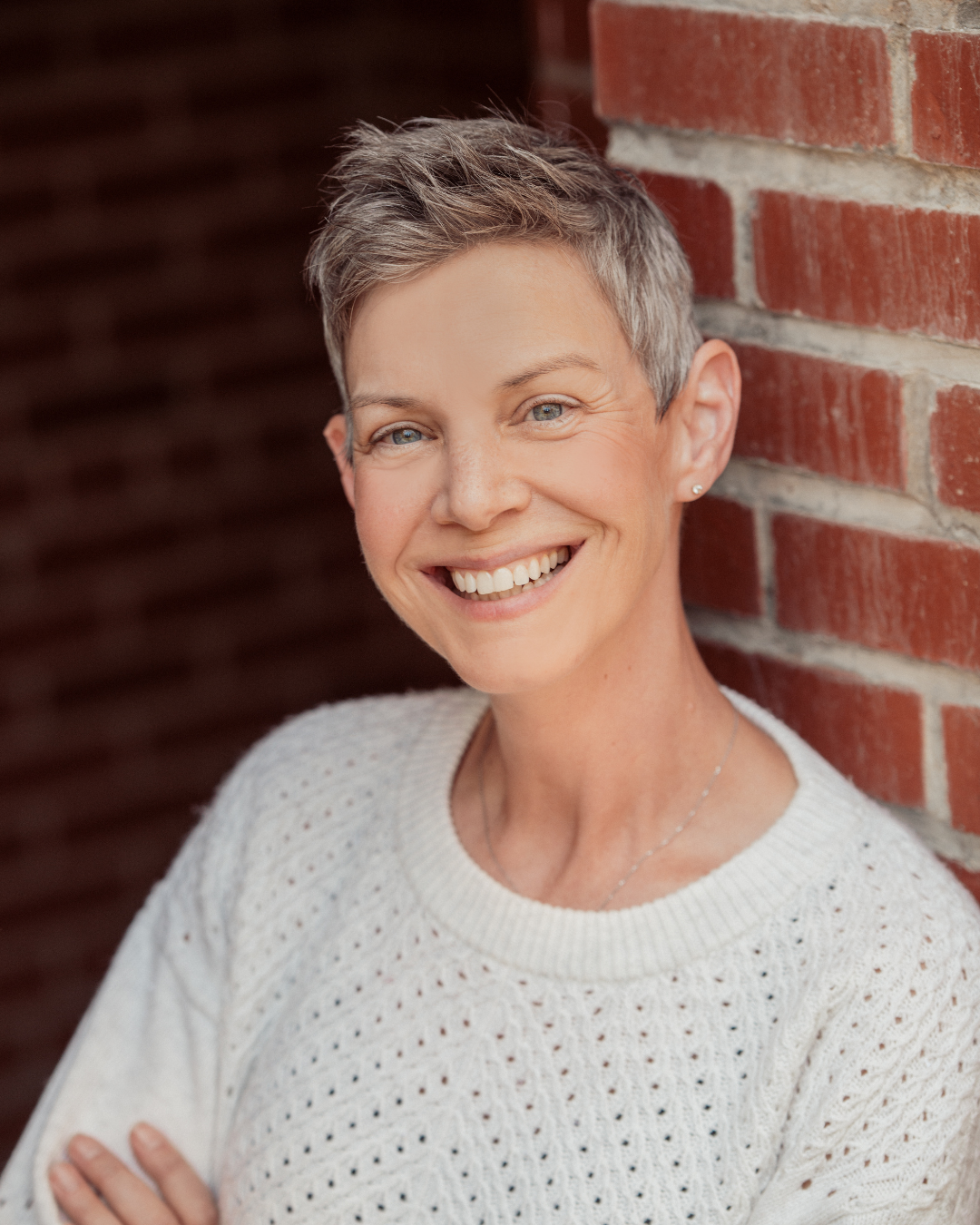 A smiling woman with short gray hair leaning against a brick wall, wearing a white knitted sweater and small earrings.