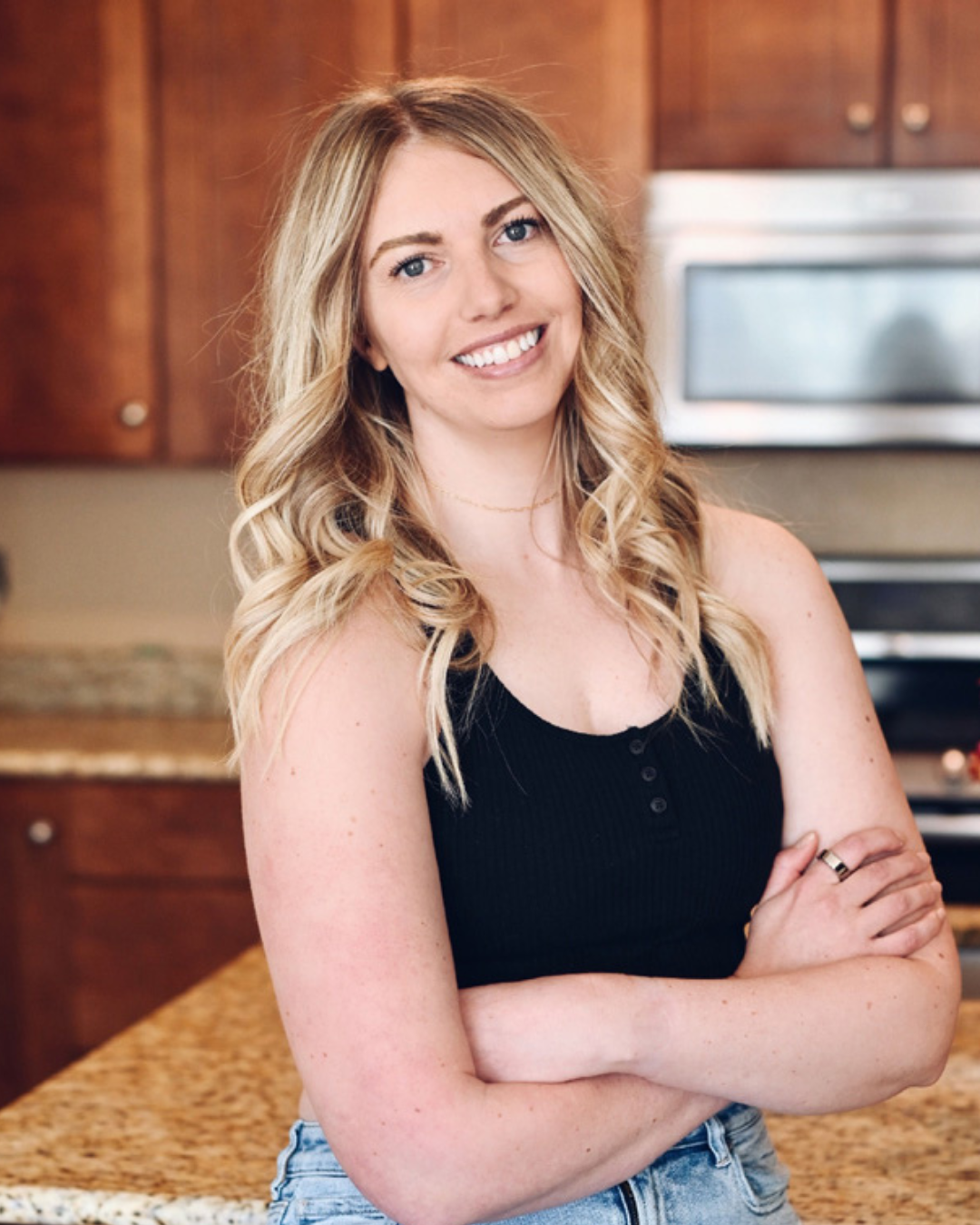 A woman with long blonde hair, wearing a black sleeveless top, smiling with arms crossed in a kitchen with wooden cabinets and a stainless steel oven in the background.
