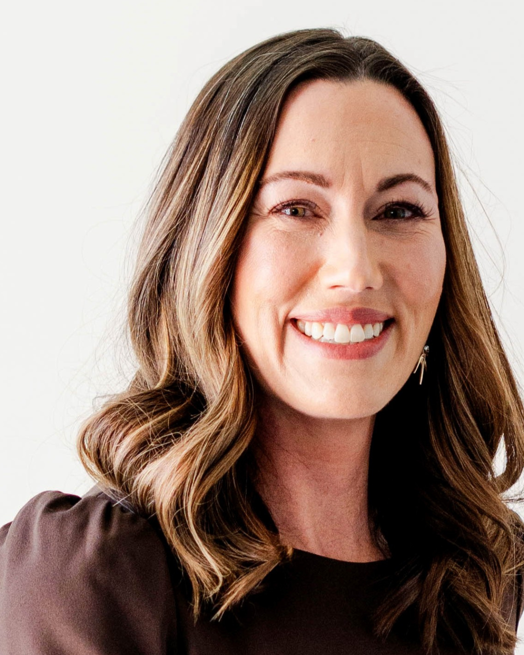 Portrait of a smiling woman with wavy brown hair, wearing a dark top, against a plain light background.