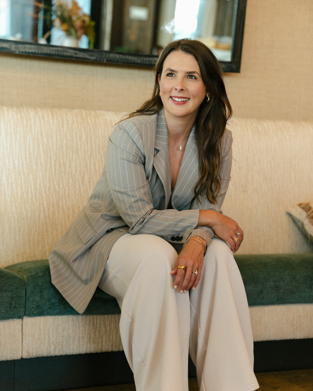 A woman with long brown hair, wearing a beige pinstripe blazer and white wide-leg pants, sits on a beige and green cushioned bench in a warmly lit room, smiling and looking to her right.