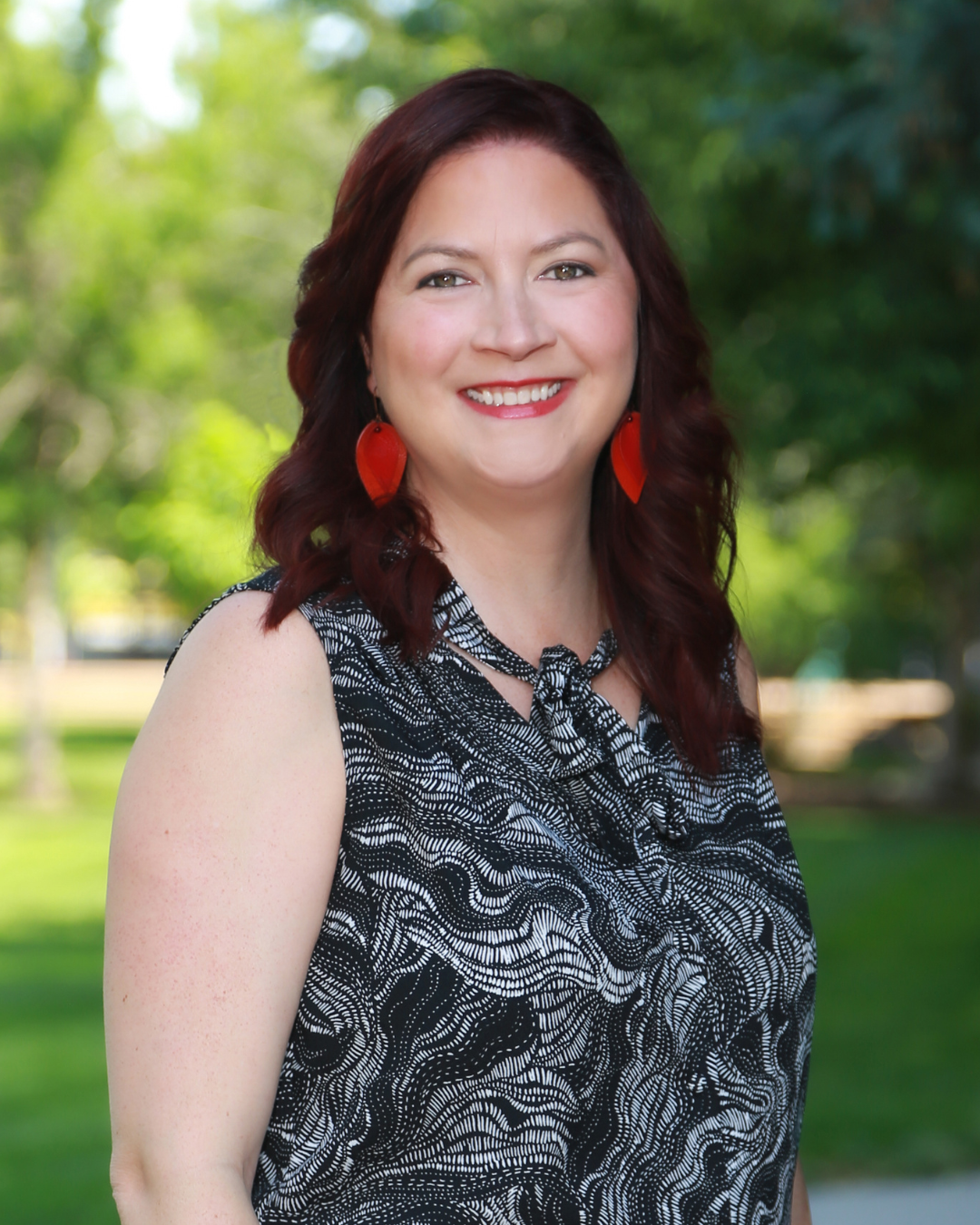 A woman with shoulder-length wavy red hair wearing red leaf-shaped earrings and a black and white patterned sleeveless top, smiling outdoors with green trees and grass in the background.