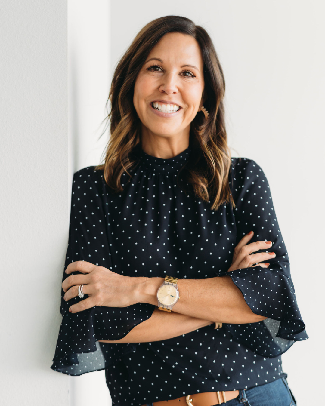 A woman with brown hair smiling, wearing a black polka dot blouse with ruffled sleeves, crossed arms, standing against a white wall.