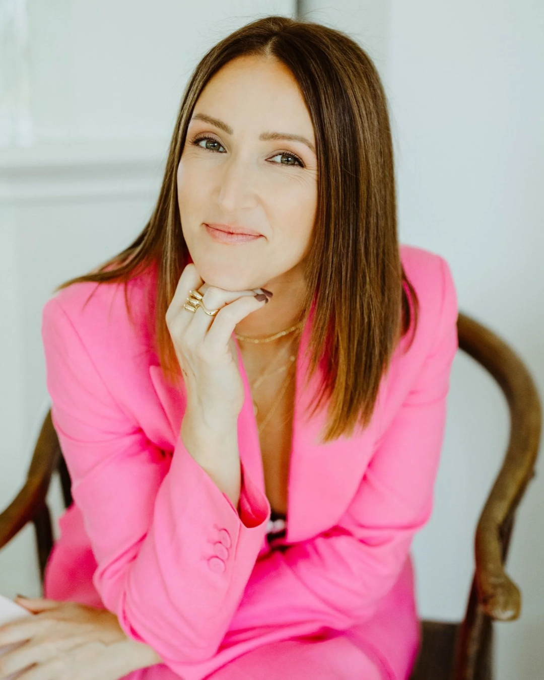 A woman with brown hair and blue eyes, wearing a pink blazer and matching lipstick, sitting on a wooden chair with a white background, smiling and resting her chin on her hand.