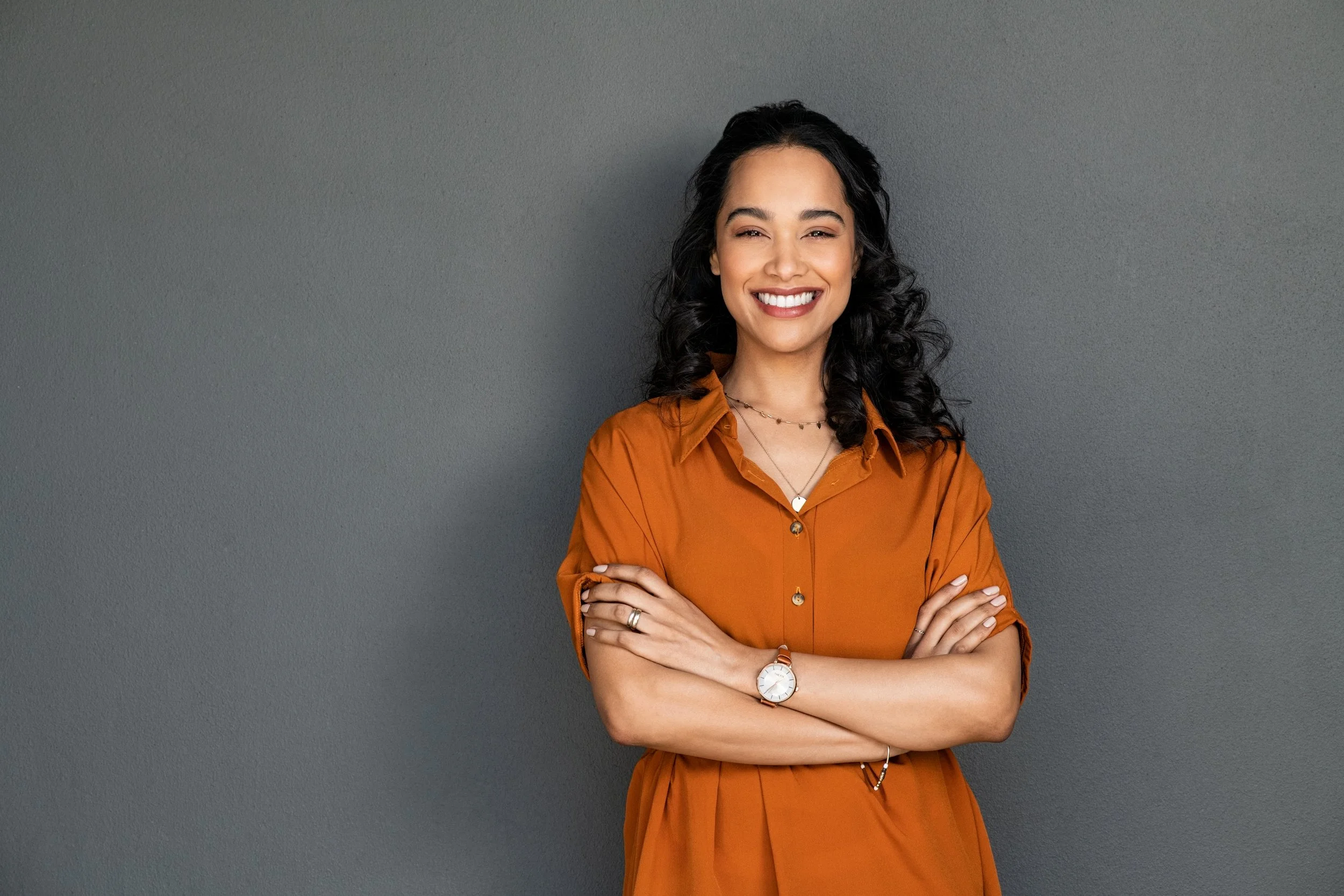 A young woman with curly black hair smiling, wearing an orange button-up shirt, crossing her arms, and standing against a gray wall.
