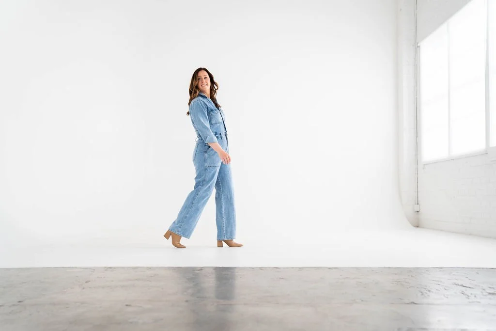 A woman in a denim jumpsuit standing in a bright, white room with large windows.