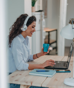 Woman wearing headphones using a laptop at a desk, holding a blue mug.