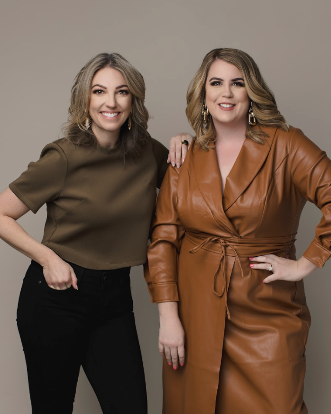 Two women with wavy hair smiling at the camera, one wearing a dark brown top and black pants, the other in a tan leather blazer dress with a waist tie, standing against a plain background.