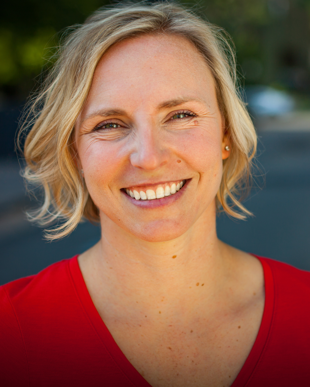 A smiling blonde woman with short hair, wearing a red top, outdoors with blurred greenery in the background.