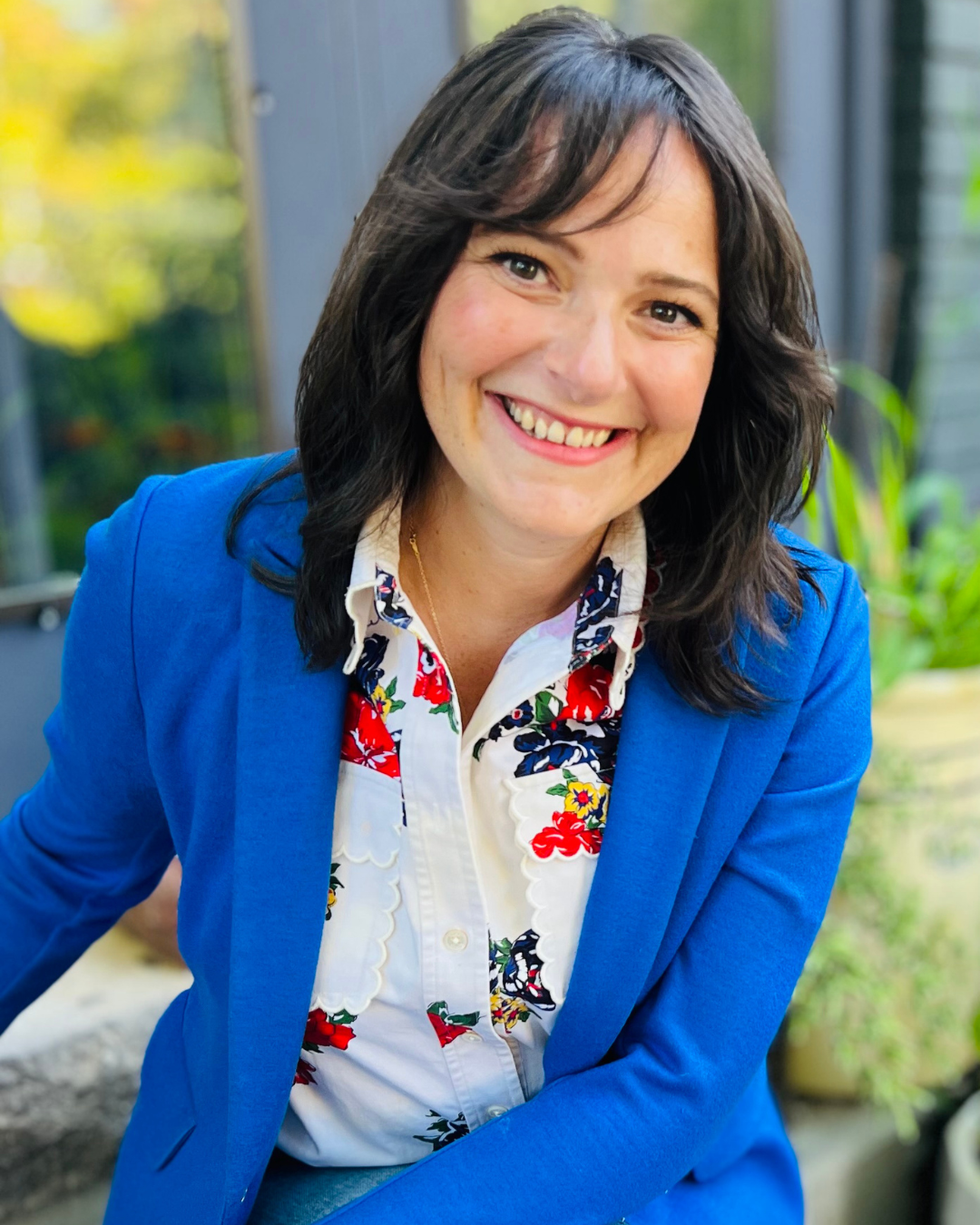 A woman smiling, with shoulder-length dark hair, wearing a bright blue blazer and a floral print blouse, sitting outdoors with blurred greenery in the background.