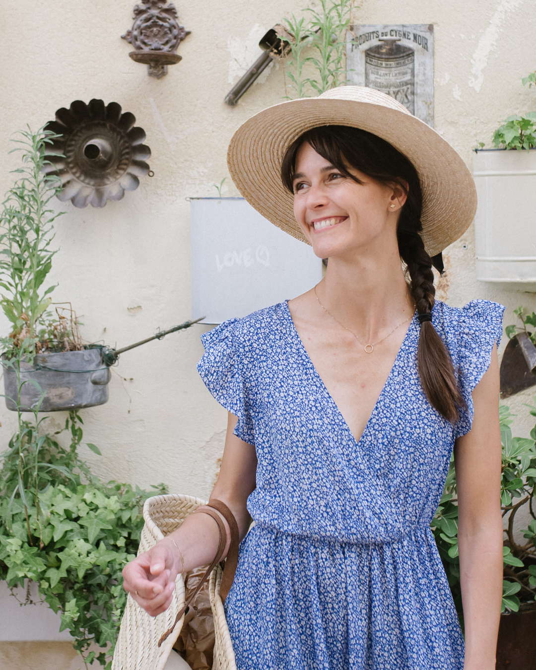 Woman with dark hair in braid, wearing a blue floral dress and a large straw hat, smiling outdoors surrounded by plants and decorative garden items.