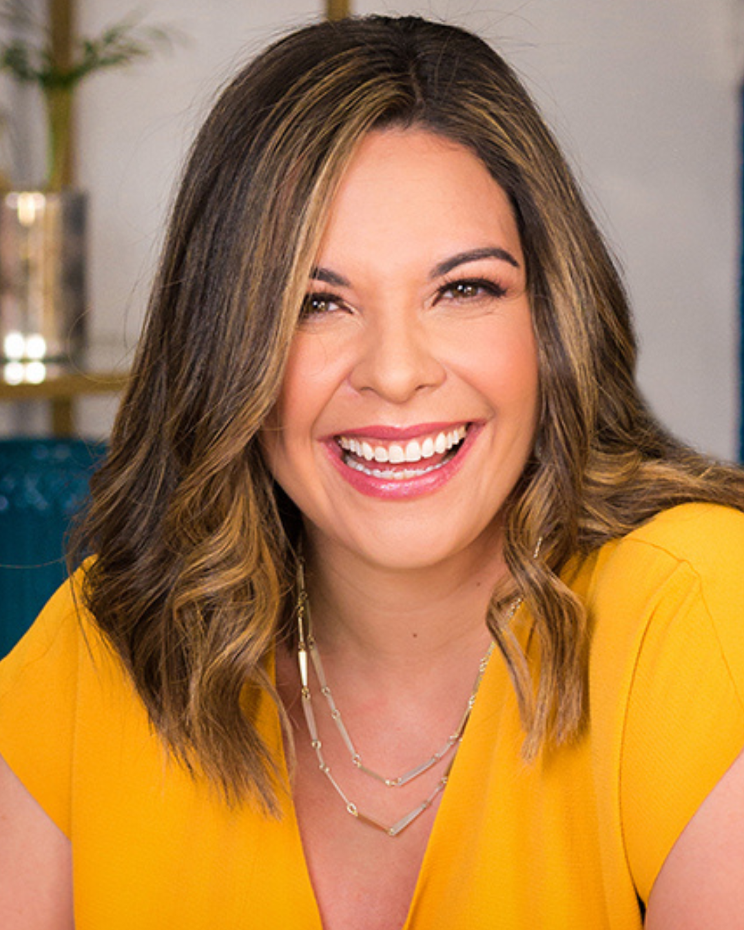 A woman with brown, wavy hair wearing a yellow top, smiling broadly showing her teeth, in an indoor setting.