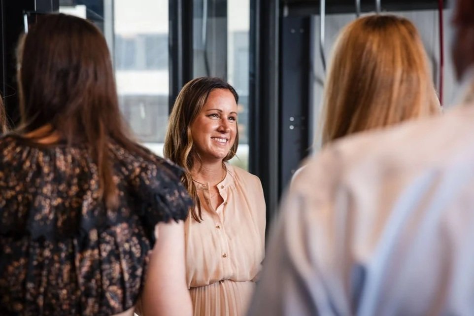 A woman smiling and talking to three other women in a social setting, with large windows in the background.