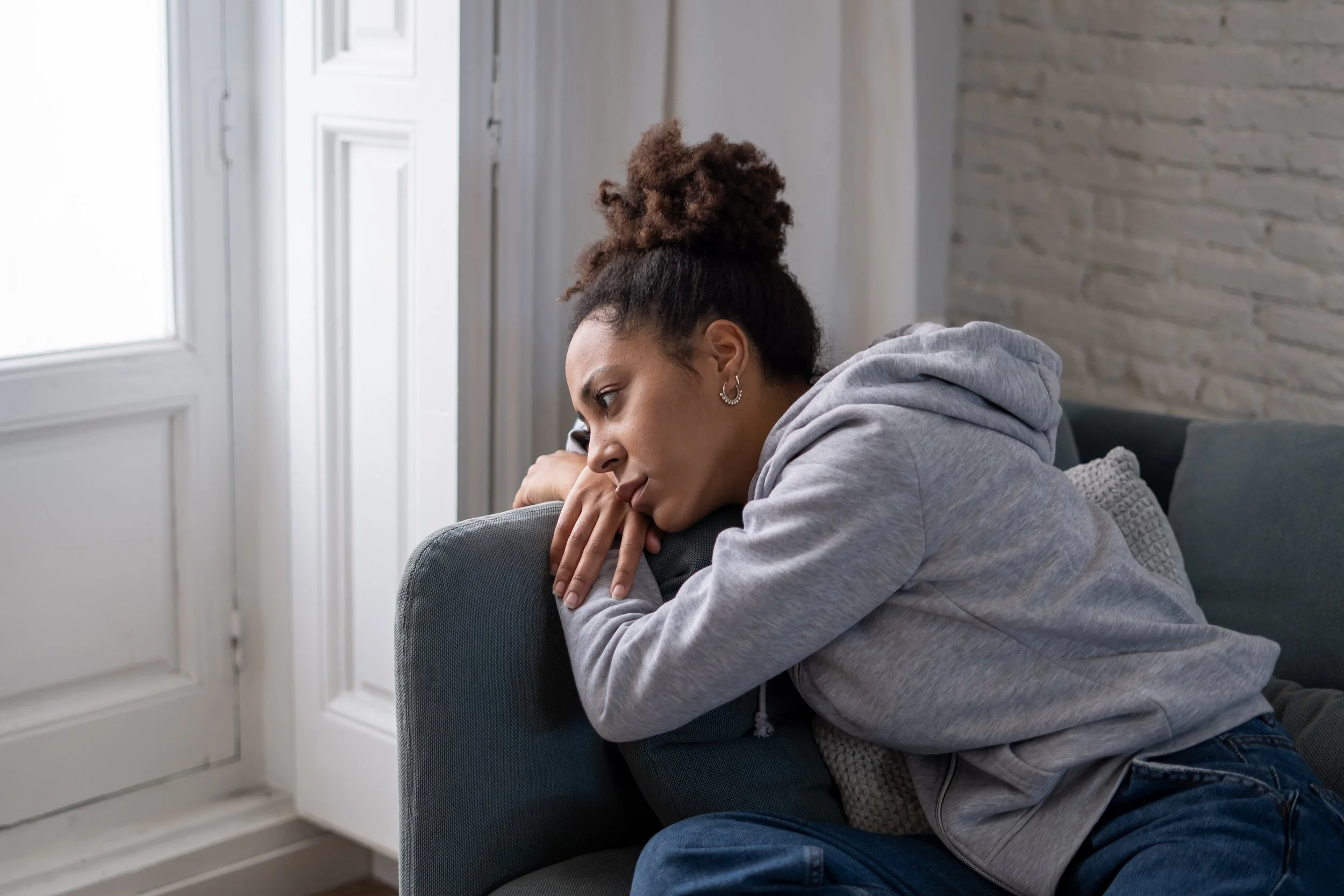 A woman with curly hair in a bun, wearing earrings and a gray hoodie, resting her head on the back of a couch near a window with white frames.