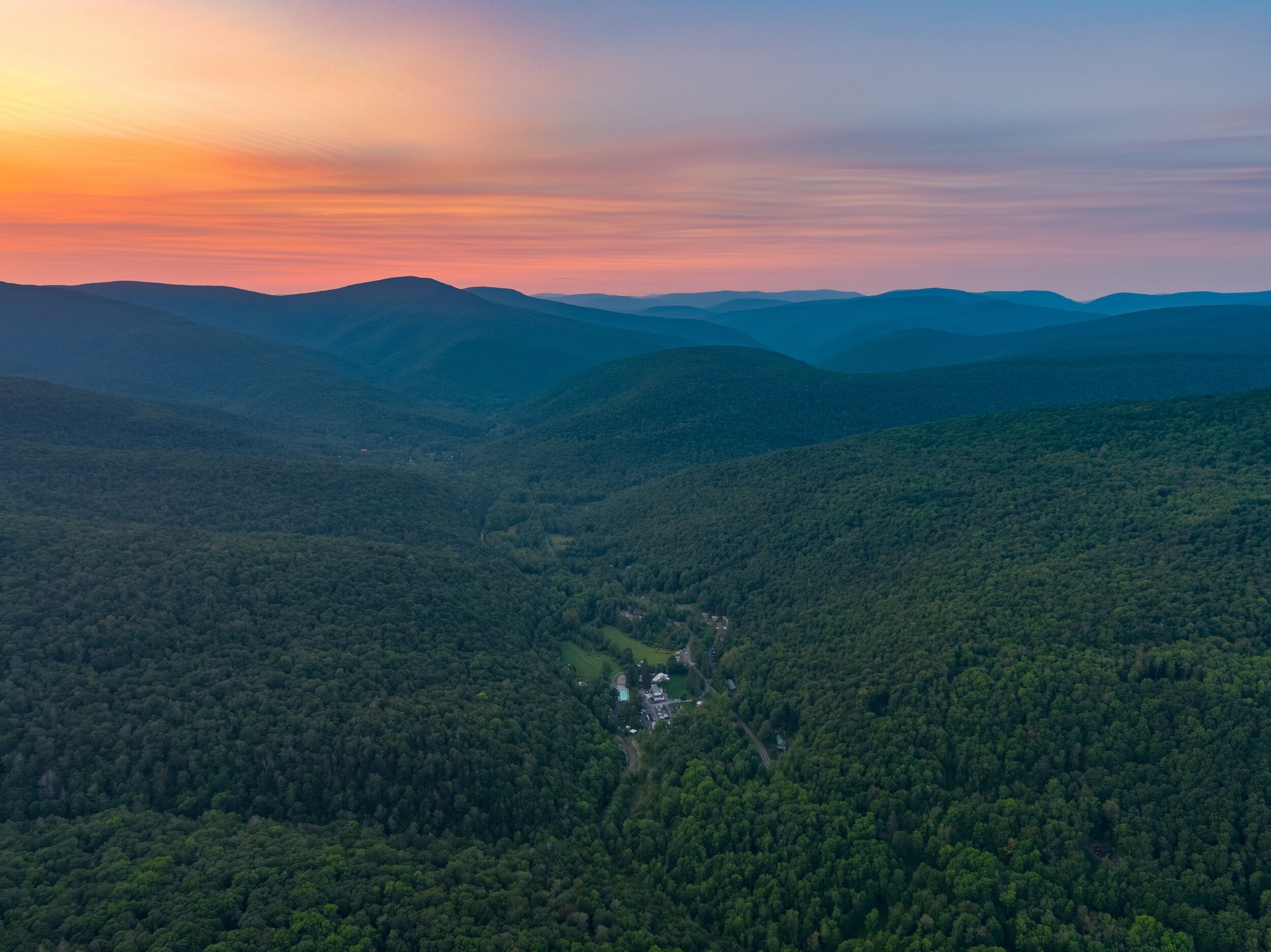 A vast, lush green mountainous landscape at sunset with a small village and winding roads amid the trees.
