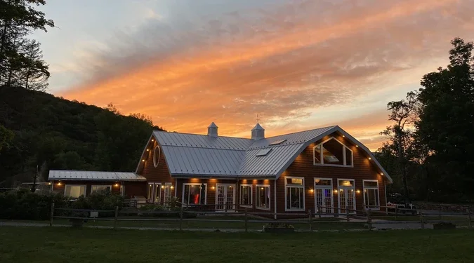 A large wooden barn with a metal roof, lit with exterior lights, at sunset with orange and pink sky, surrounded by trees and a grassy area.