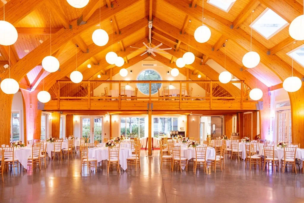 A spacious banquet hall with wooden beams and walls, decorated with hanging white paper lanterns, set up with round tables covered in white tablecloths, floral centerpieces, and wooden chairs, with large windows allowing natural light.