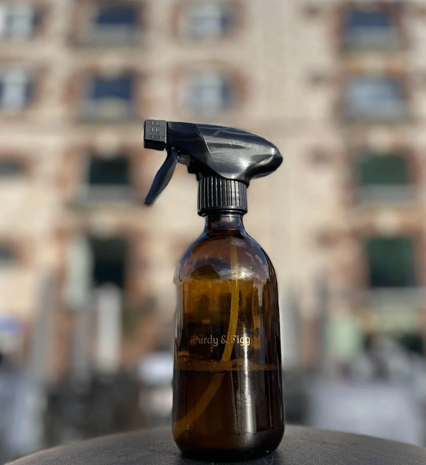 A brown glass spray bottle with a black spray nozzle and trigger, labeled 'Purdy & Figg', standing outdoors with a blurred building in the background. Eco Friendly products which smell amazing.