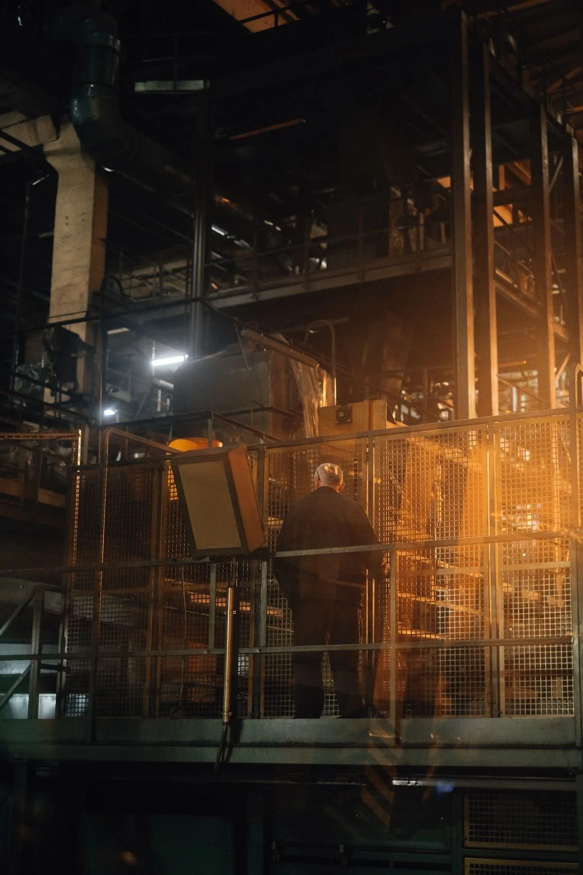 An older man with gray hair, dressed in black, inside a factory or industrial setting, surrounded by metal scaffolding and machinery, illuminated by orange safety lights.