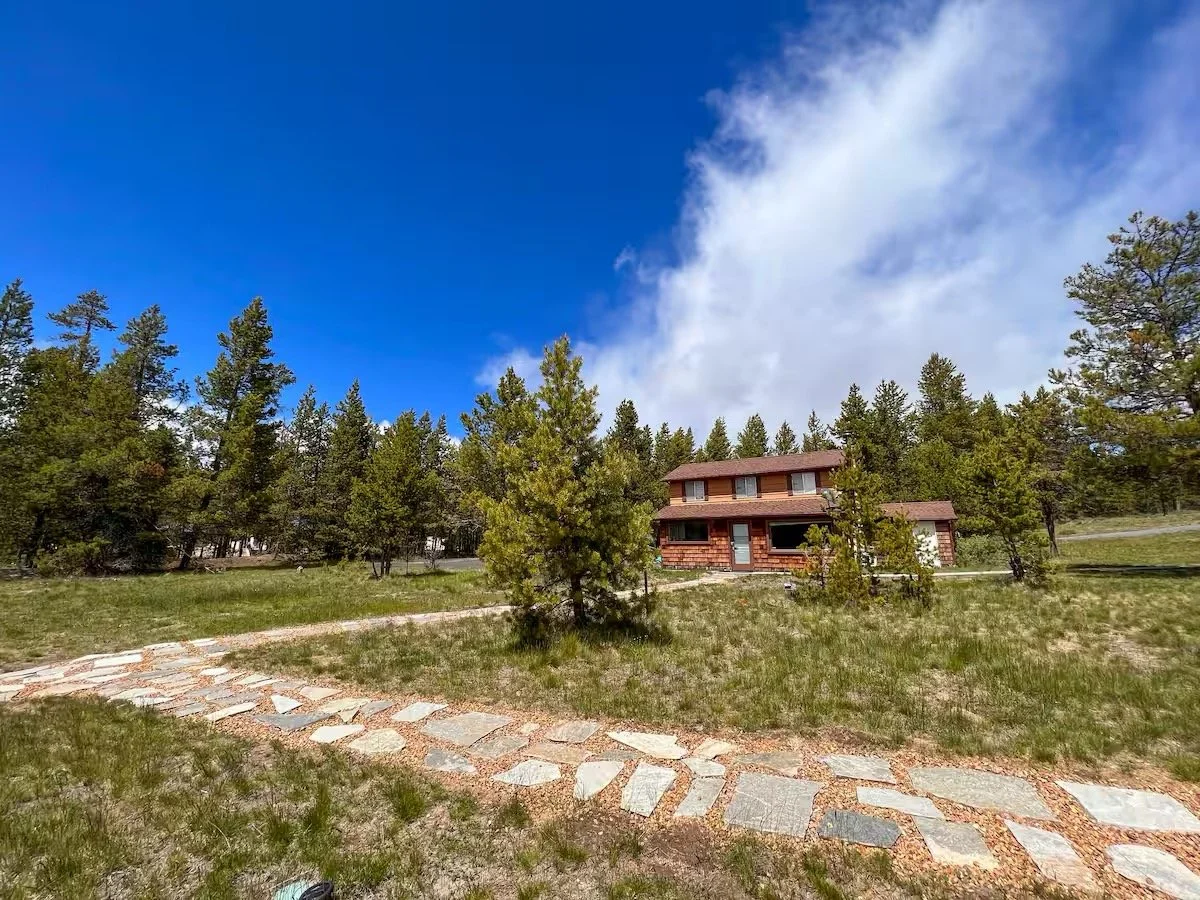 A rustic two-story wooden house in a forested area with tall pine trees, a stone pathway leading to the house, and a partly cloudy blue sky overhead.