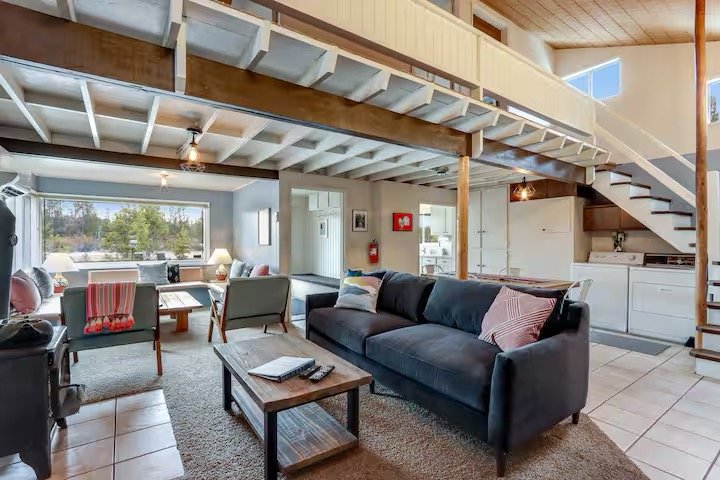 Living room and kitchen area with a vaulted ceiling, staircase, large window, and modern furnishings.