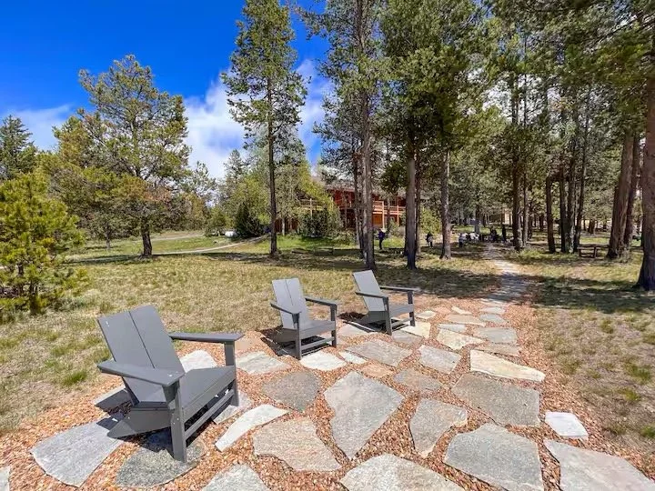 Three gray outdoor chairs arranged on a stone pathway in a wooded park with tall trees and a building in the background.
