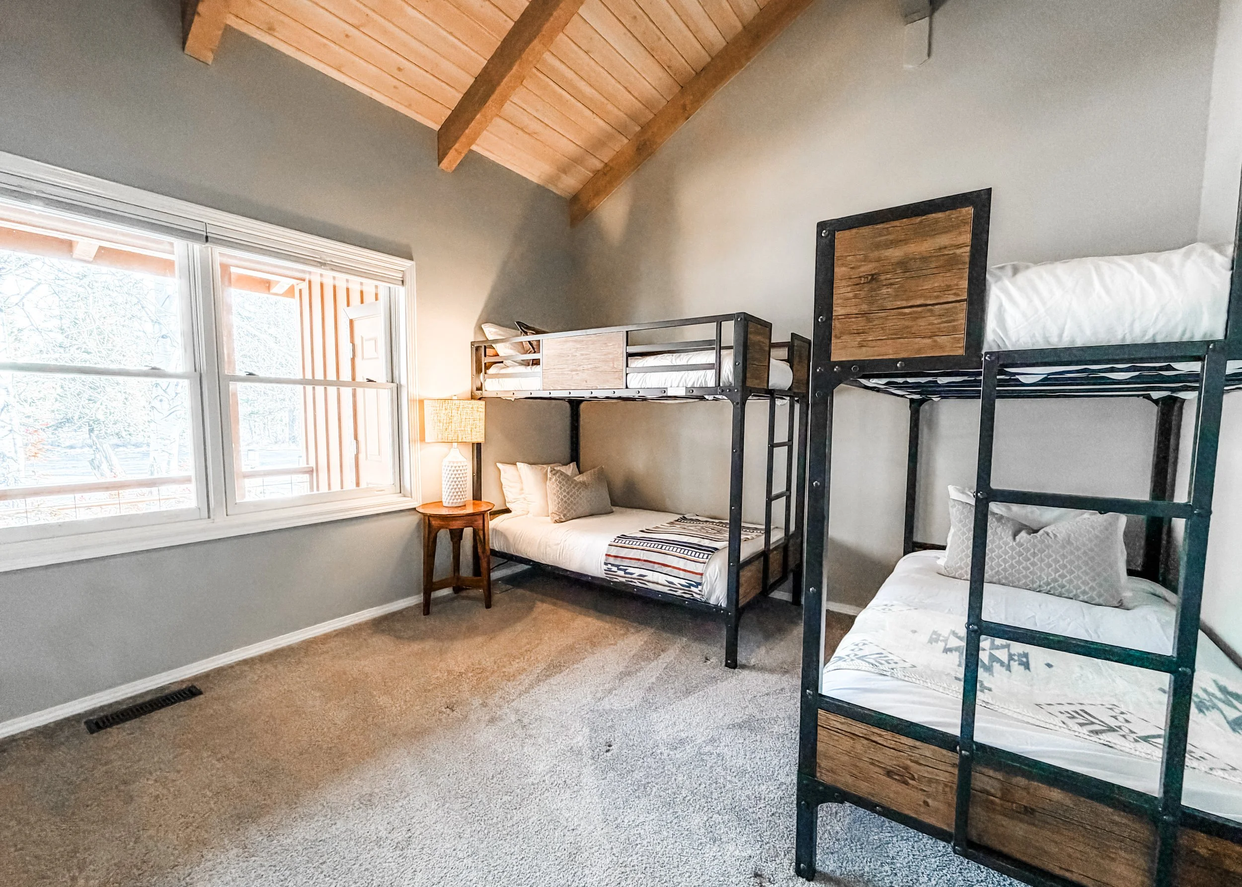 A bedroom with a large window, a small wooden nightstand with a white lamp, a bunk bed with black metal frames and wooden accents, beige carpeted floor, grey walls, and a wooden ceiling with exposed beams.