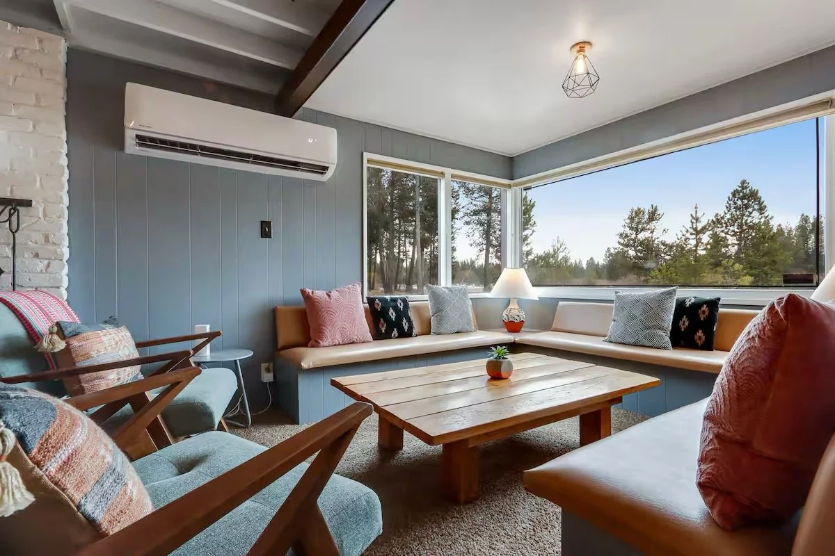 Living room with large corner windows showing trees, gray walls, a wooden coffee table, a white sofa with multiple decorative pillows, two armchairs, a table lamp, and an air conditioning unit.