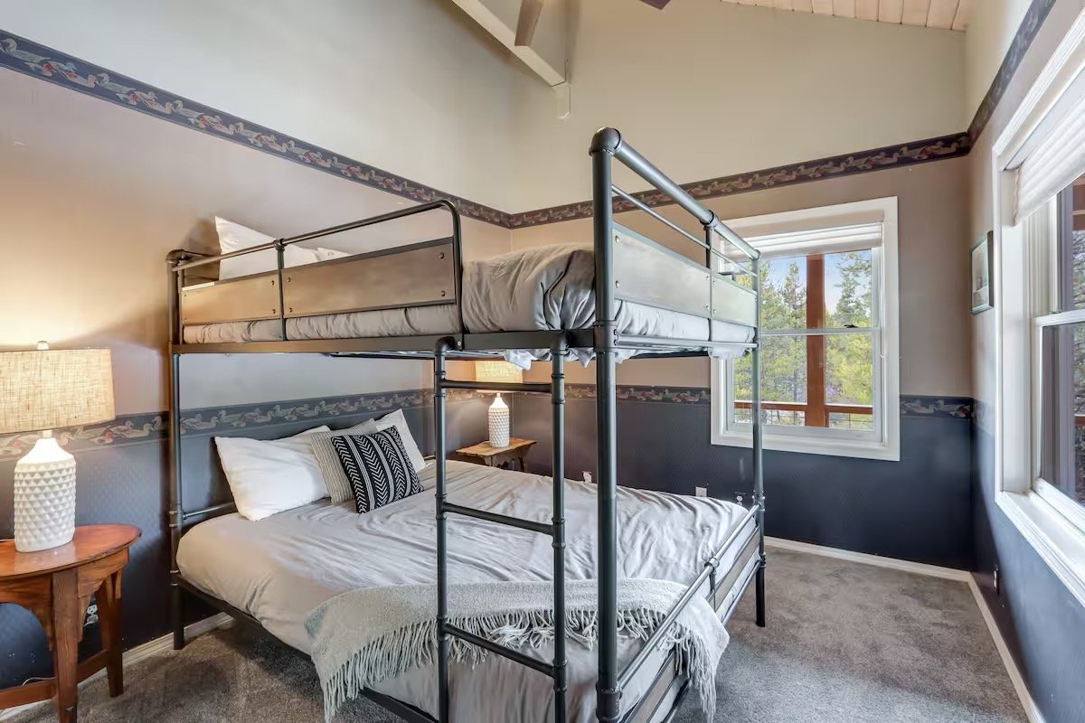 A bedroom with a metal bunk bed, a large window, a nightstand with a lamp, and two pillows on the bed.