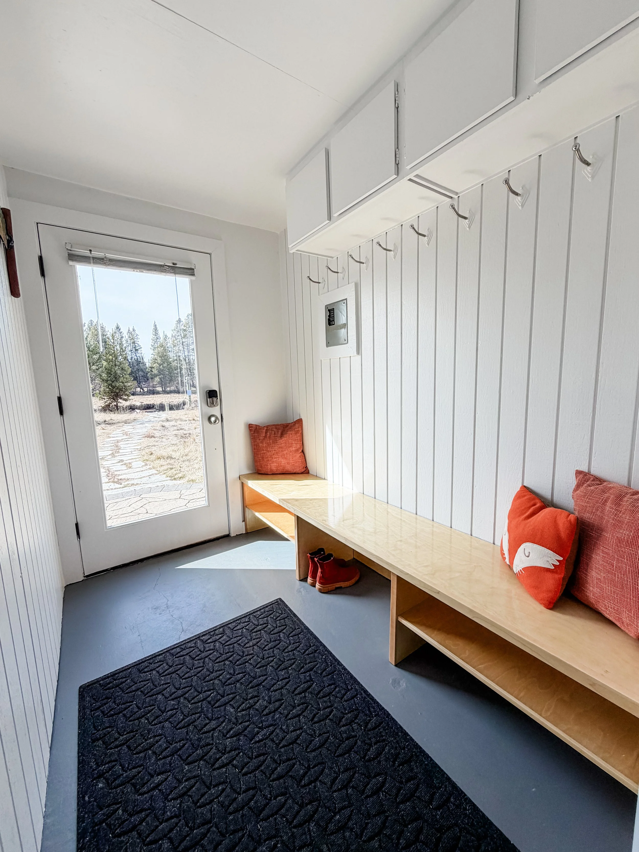 Small entryway with white paneled walls, a glass door leading outside, a wooden bench with orange pillows and red shoes underneath, a black textured mat on a concrete floor, and hooks with folded items hanging above the bench.