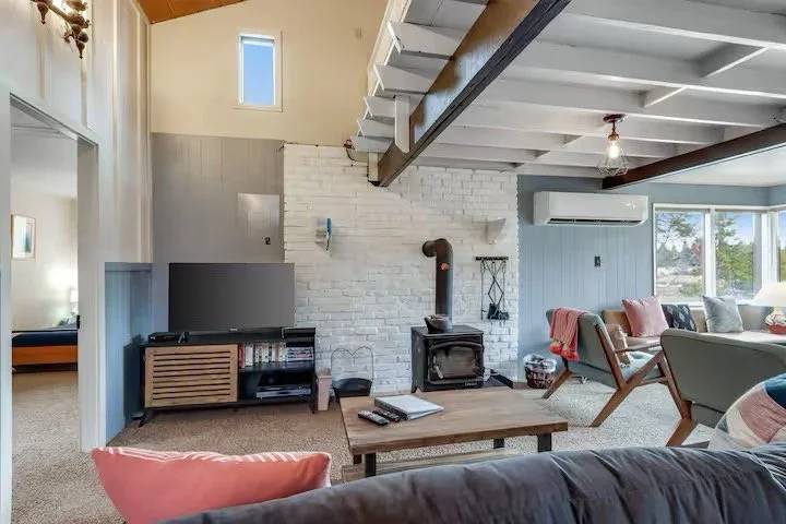 Living room with white brick fireplace, wood stove, and large windows with view of trees, furnished with colorful chairs, a wooden coffee table, and TV.