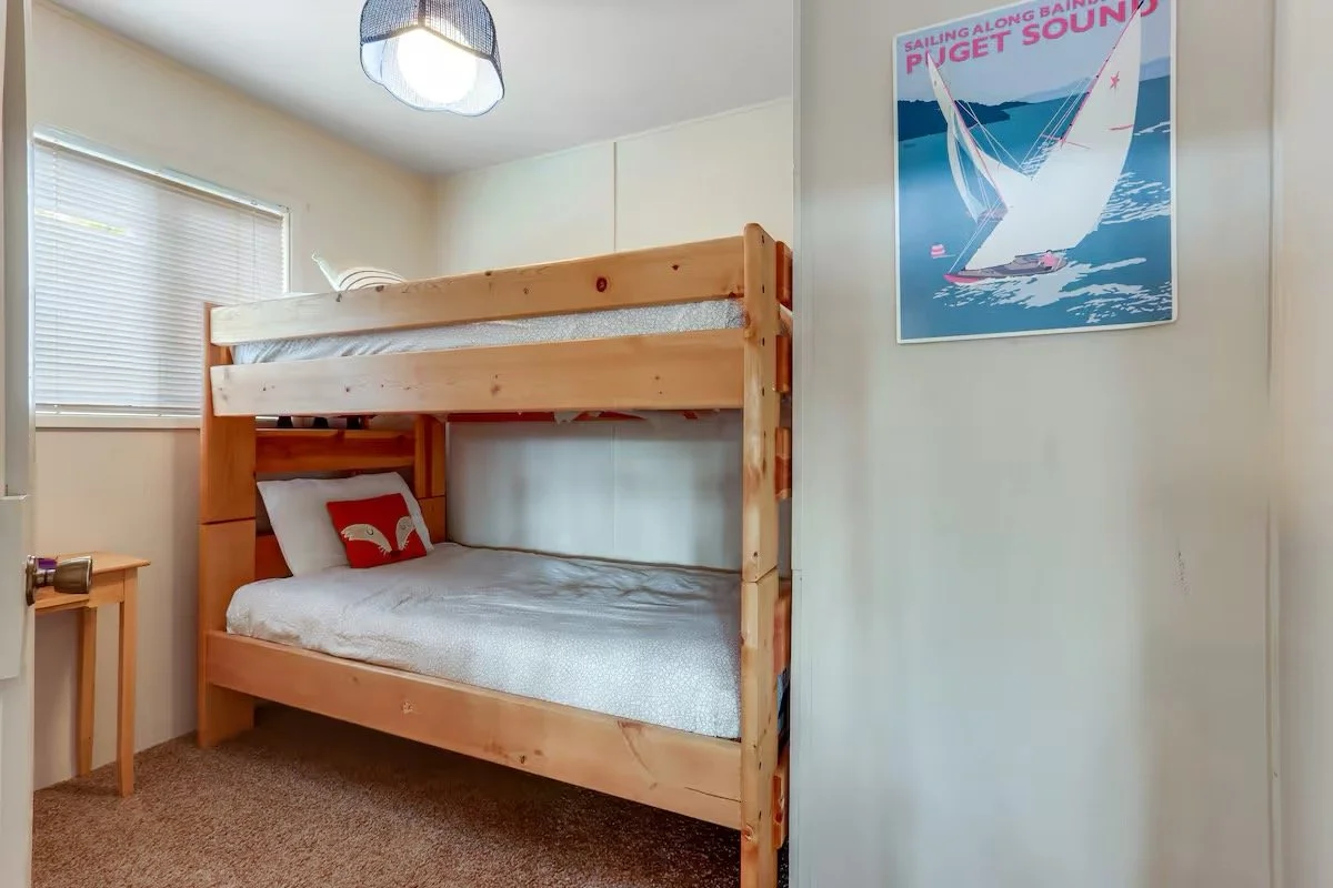 A small bedroom with a wooden bunk bed, a window with blinds, a small wooden side table, and a colorful sailing-themed poster on the wall.