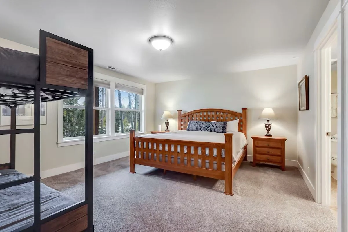 A bedroom with a wooden bed, two nightstands with lamps, large windows, and a bunk bed on the left, with beige carpet and white walls.