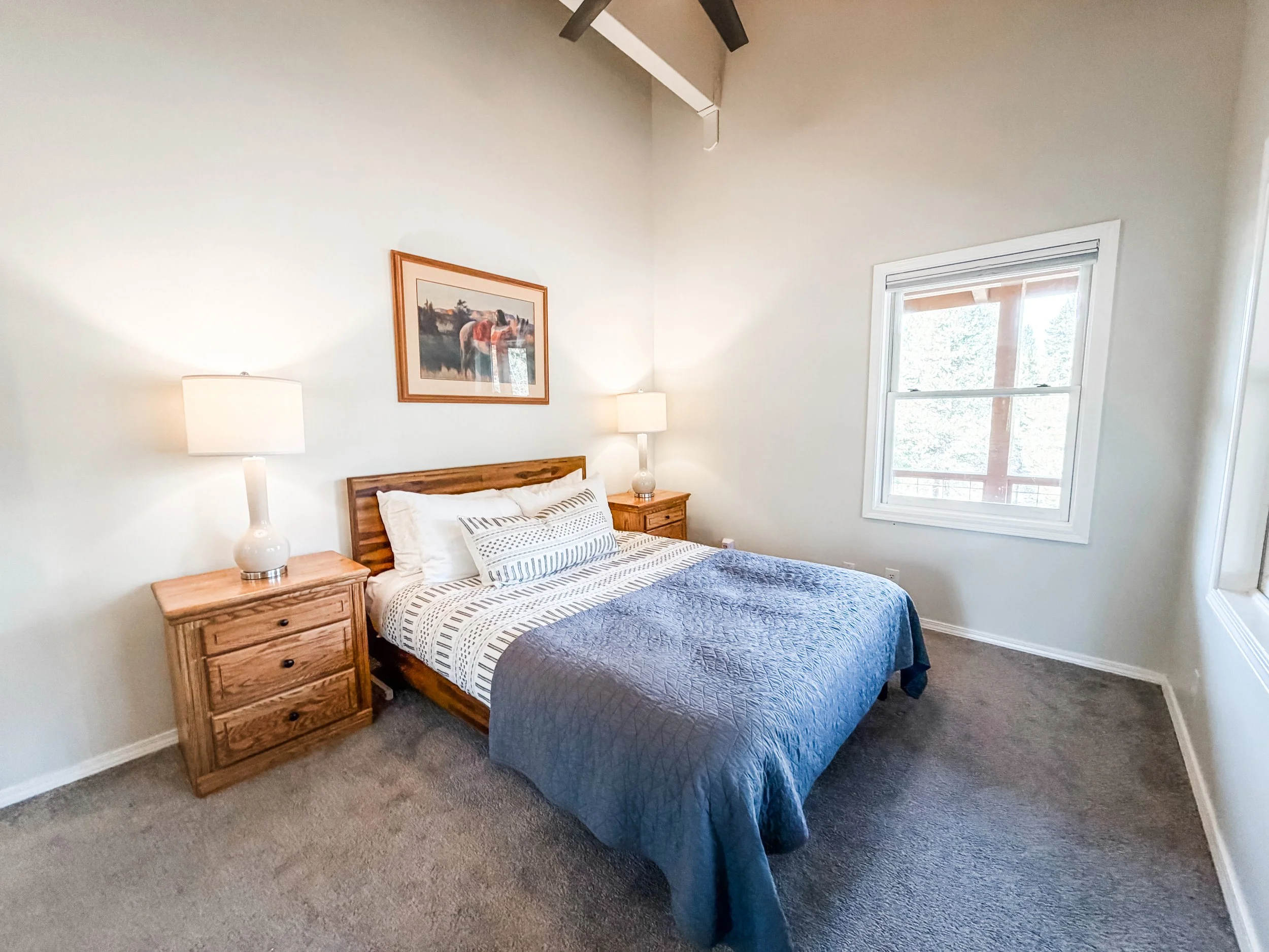 A bedroom with a wooden bed frame, striped bedding, a blue quilt, and pillows. Two wooden nightstands with matching lamps and a window with white trim, beige carpet, and a picture hanging on the wall above the bed.