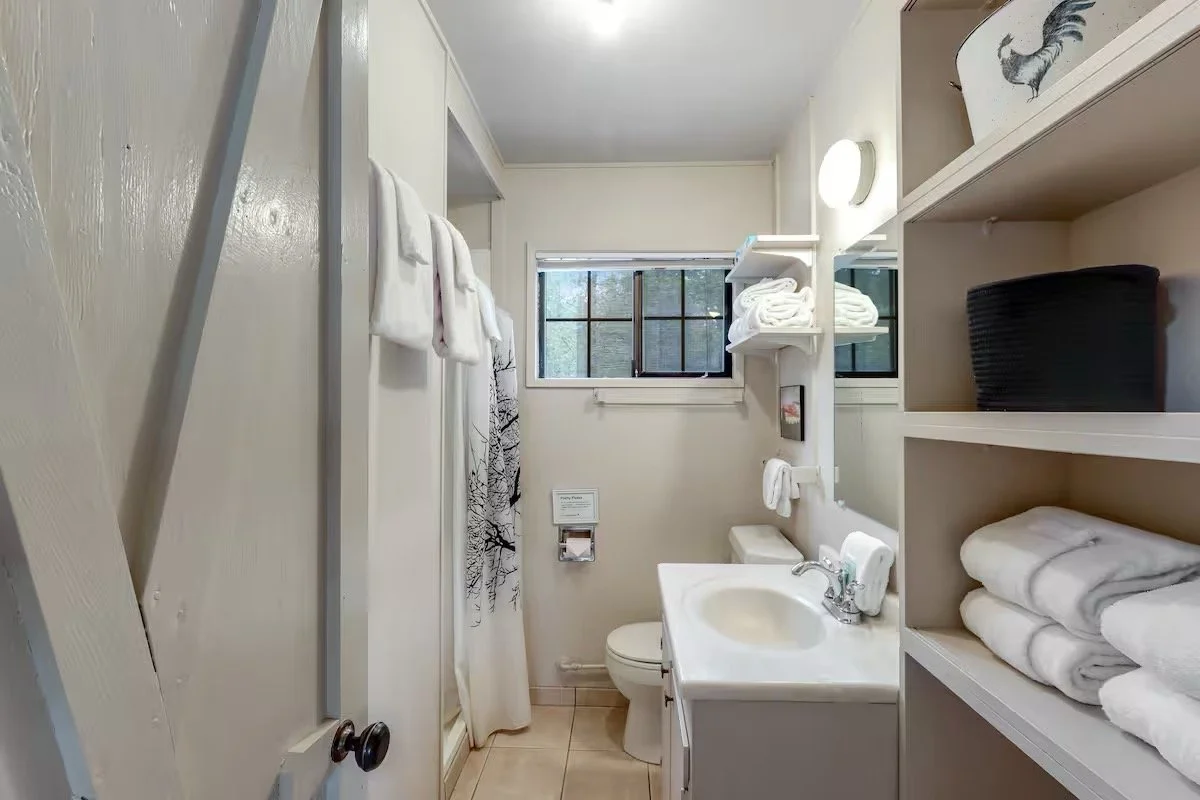 Small bathroom with white walls and beige tile floor, featuring a window with blinds, a white toilet, a white sink with a mirror, shelves with folded towels and black storage basket, and a shower with a curtain and towel hanging on the door.