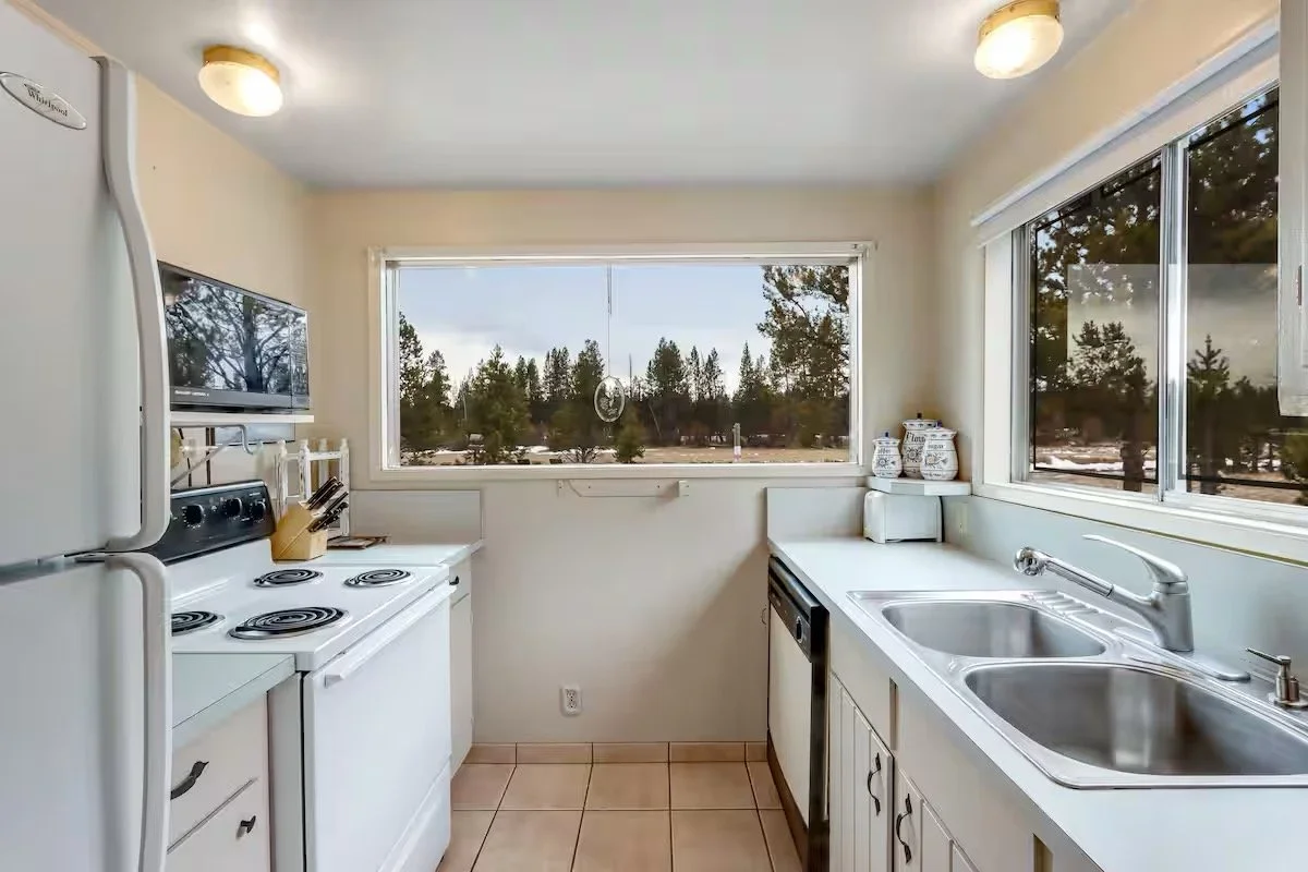 Kitchen with white countertops, double sink, window view of trees, white stove, and small appliances.
