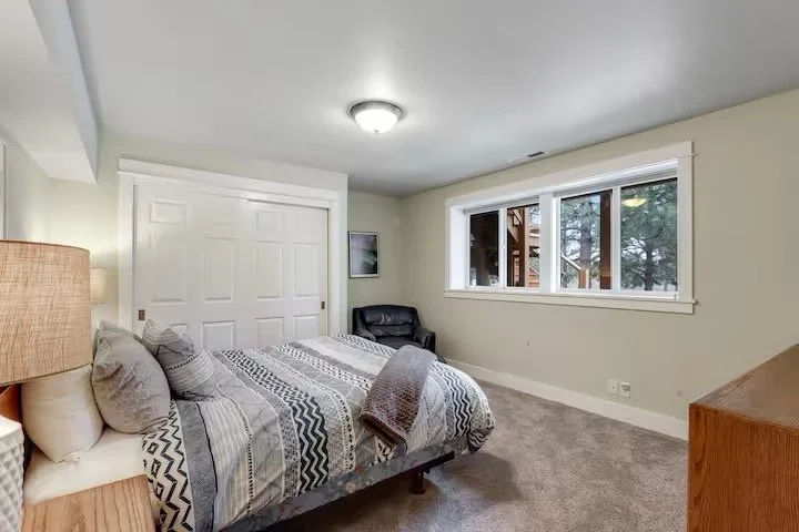A bedroom with a bed, pillows, and a blanket, a black chair near the window, a framed picture on the wall, white closet doors, and a dresser, with a view of trees outside through the window.