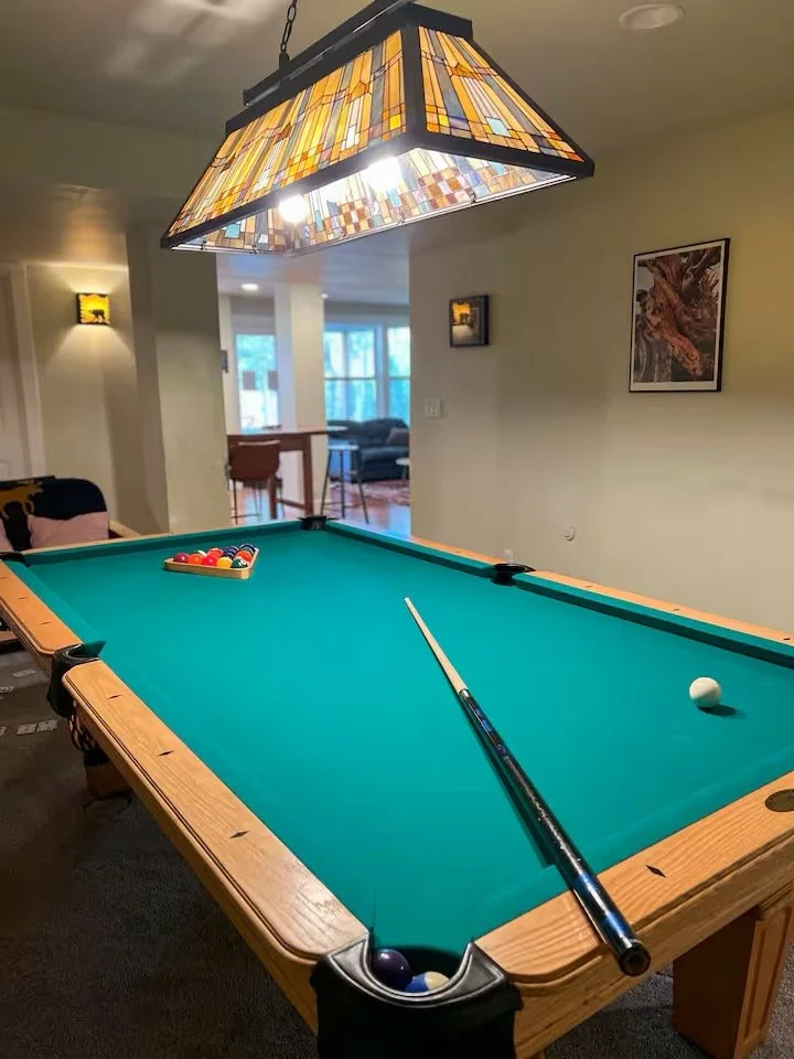 A pool table with a green felt surface, positioned under a stained glass ceiling light fixture, in a cozy living room setting.