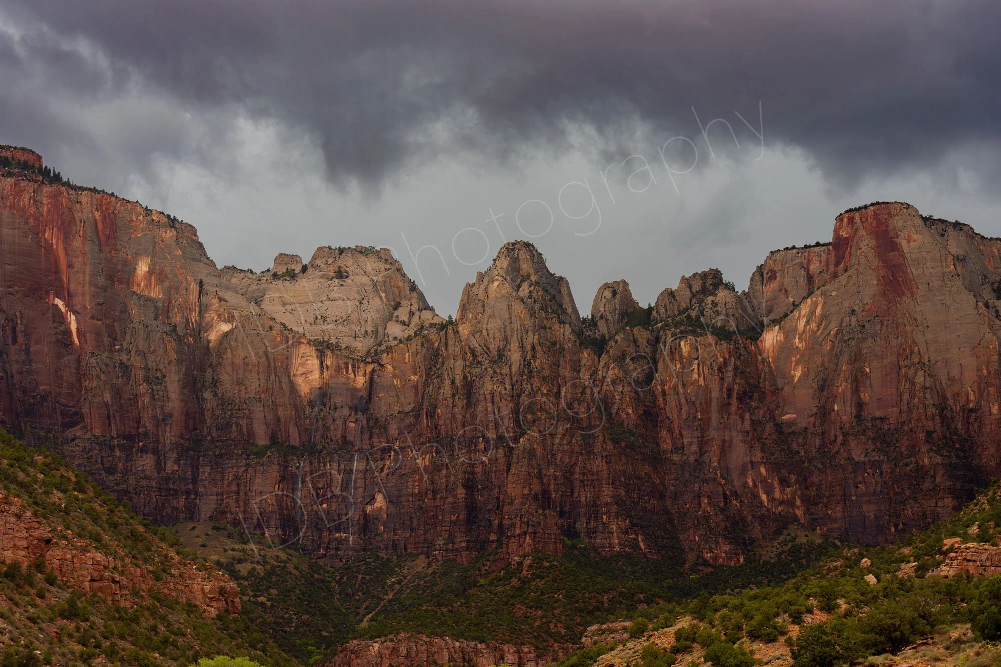 A zoomed in image of the beautiful brown, red and grey irregular cliffs below a stormy sky at Zion National Park, Utah with shrubby green foreground.