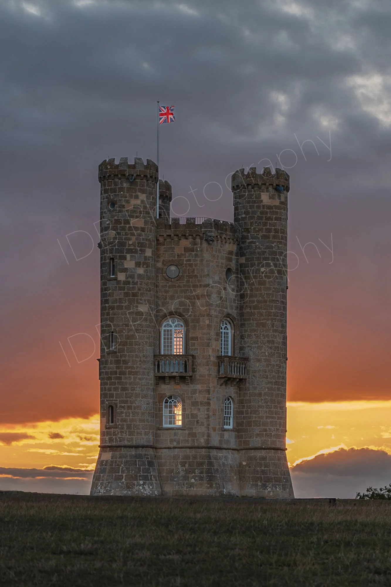 Broadway Tower, Cotswolds at sunset, the setting sun is behind the tower illuminating the windows with an orange glow, dark cloudy sky.